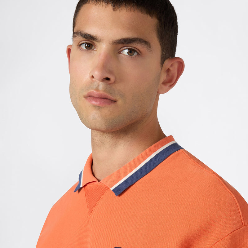 A young man with short dark hair models the Champion Men’s Knitted Polo Neck Sweatshirt in orange with blue and white trim, showcasing contemporary men’s sportswear against a plain light background.