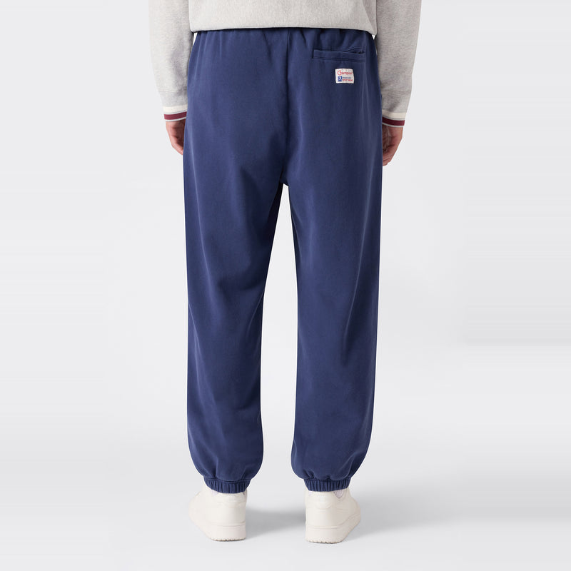 A person is wearing Champion Men’s Knitted Elastic Cuff Pants in navy blue, which feature a relaxed fit and a small label on the back, paired with white shoes and a light gray top with maroon-trimmed sleeves, standing against a plain light background.