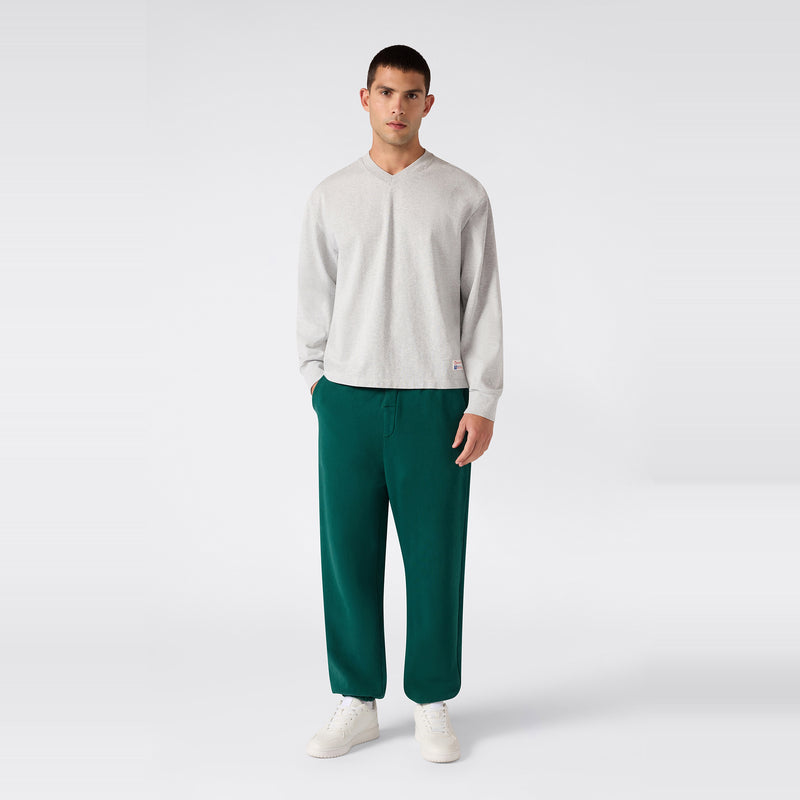A man stands facing forward, wearing a light gray long-sleeve shirt, Champion Men’s Knitted Elastic Cuff Pants in teal green, and white sneakers, against a plain white background.