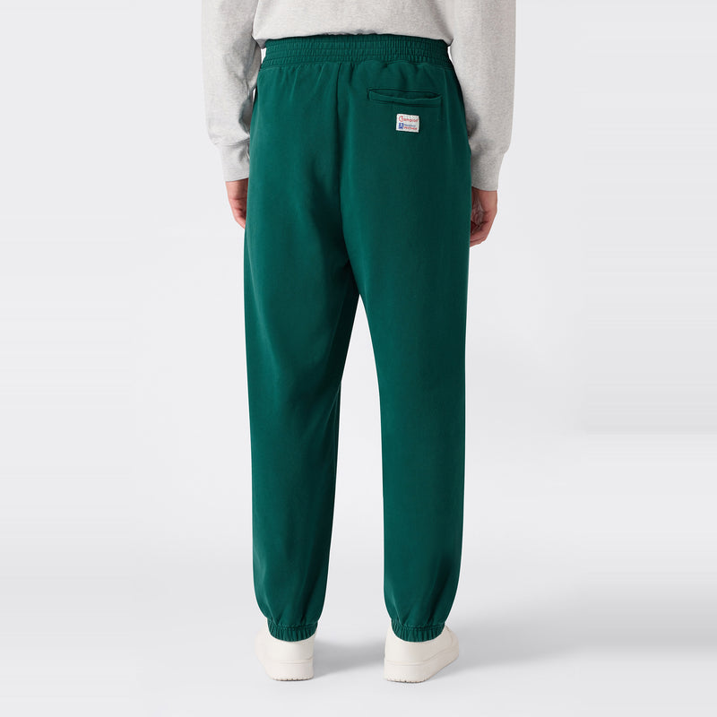 A person stands with their back to the camera wearing green Men’s Knitted Elastic Cuff Pants by Champion and a light gray sweatshirt. The pants, inspired by modern Ametora trousers, feature an elastic waistband and cuffs, a back pocket, and a small rectangular patch above the pocket.
