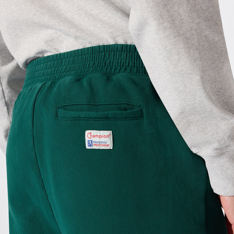 A person is wearing green Champion Men’s Knitted Elastic Cuff Pants featuring a back pocket and a white Champion logo patch, paired with a light grey long-sleeve shirt.