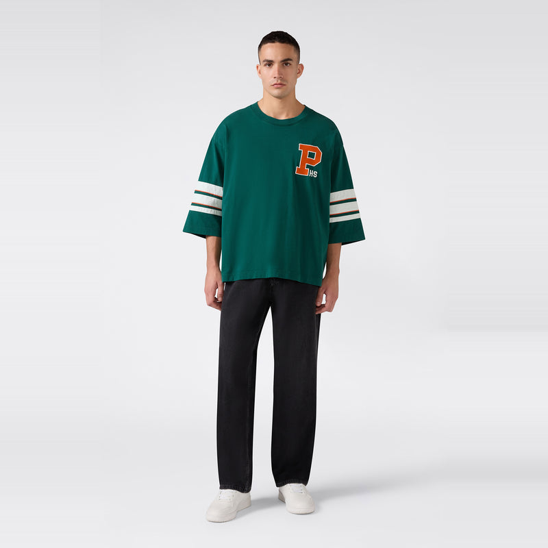 A man stands against a plain white background, wearing the Champion Men’s Knitted Crewneck T-Shirt—a relaxed fit green shirt featuring white stripes on the sleeves and a large orange letter P on the chest—paired with black pants and white sneakers.