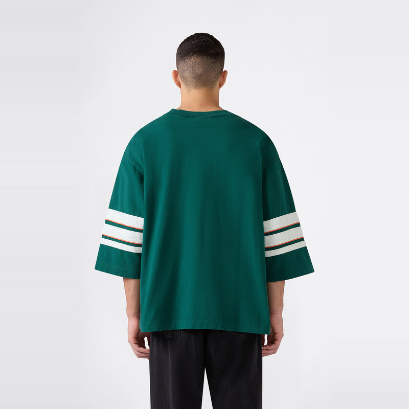 A person stands with their back to the camera, wearing the Champion Men’s Knitted Crewneck T-Shirt in a relaxed fit with wide green sleeves featuring white stripes, paired with black pants against a plain light background.