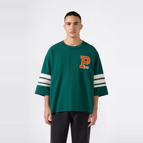 A person stands facing forward wearing the Champion Men’s Knitted Crewneck T-Shirt in green, featuring a relaxed oversized fit, white stripes on the sleeves, and a large orange “P HS” patch on the left chest. The iconic design is inspired by the classic Champion Reverse Weave t-shirt. The background is plain white.