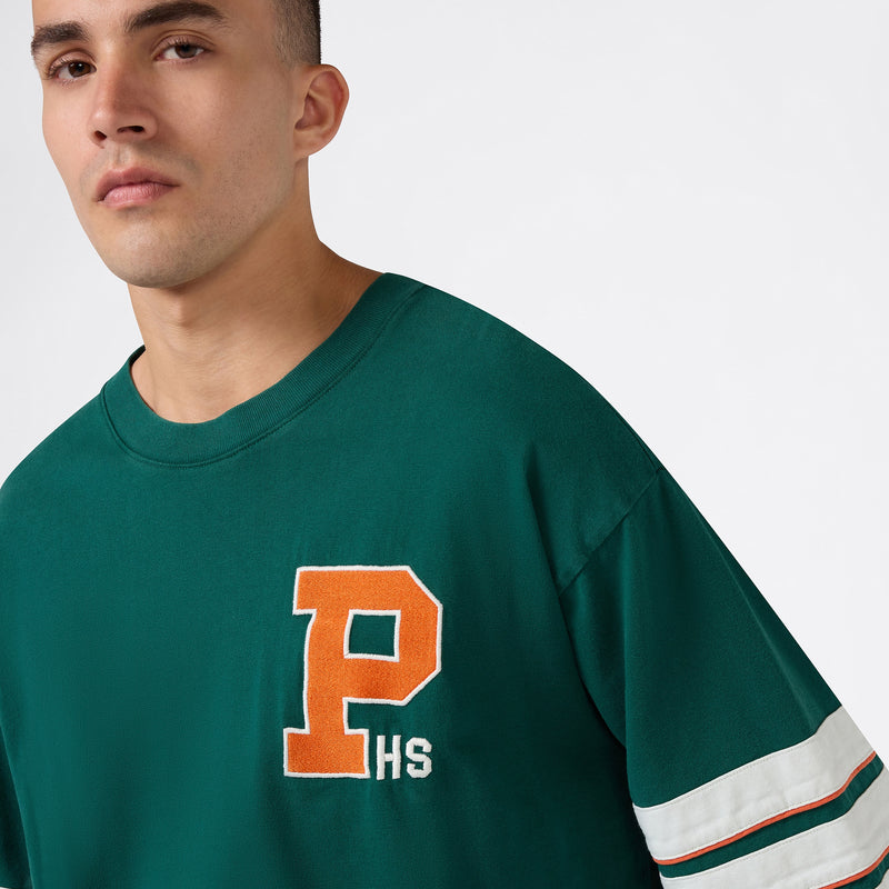 A person wearing a green Champion Men’s Knitted Crewneck T-Shirt is looking at the camera against a plain background. This crewneck features a relaxed fit for everyday comfort, with white sleeve stripes and an orange letter P above HS on the front.