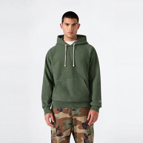 A man stands facing forward against a plain white background, wearing a green Champion Men’s Relaxed Fit Hooded Sweatshirt with drawstrings and camouflage pants.