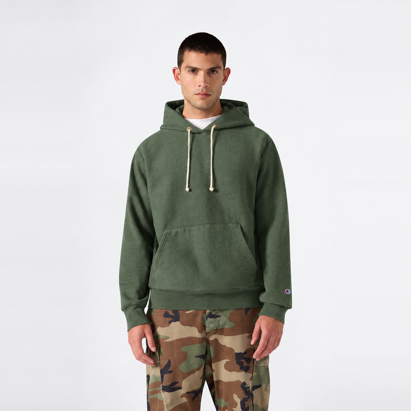 A man stands facing forward against a plain white background, wearing a green Champion Men’s Relaxed Fit Hooded Sweatshirt with drawstrings and camouflage pants.
