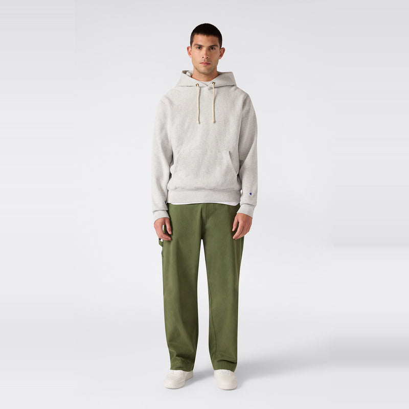 A man stands facing forward against a plain white background, wearing a relaxed fit Men’s Knitted Hooded Sweatshirt by Champion in light gray, paired with olive green pants and white shoes.
