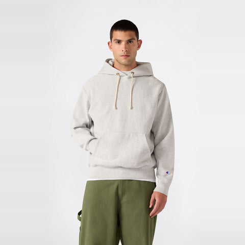 A person stands against a plain background wearing a light gray Champion Men’s Knitted Hooded Sweatshirt with drawstrings, paired with olive green pants. Looking directly at the camera with one hand in the hoodie pocket, he showcases this classic fleece designed for durability and comfort.