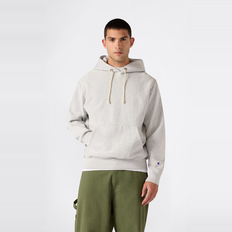 A person stands against a plain background wearing a light gray Champion Men’s Knitted Hooded Sweatshirt with drawstrings, paired with olive green pants. Looking directly at the camera with one hand in the hoodie pocket, he showcases this classic fleece designed for durability and comfort.