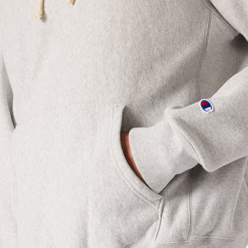 A person wearing a light gray Champion Men’s Knitted Hooded Sweatshirt with one hand inside the front pocket. The sleeve features a small embroidered Champion logo near the wrist, accentuating the classic design of this men’s fleece.
