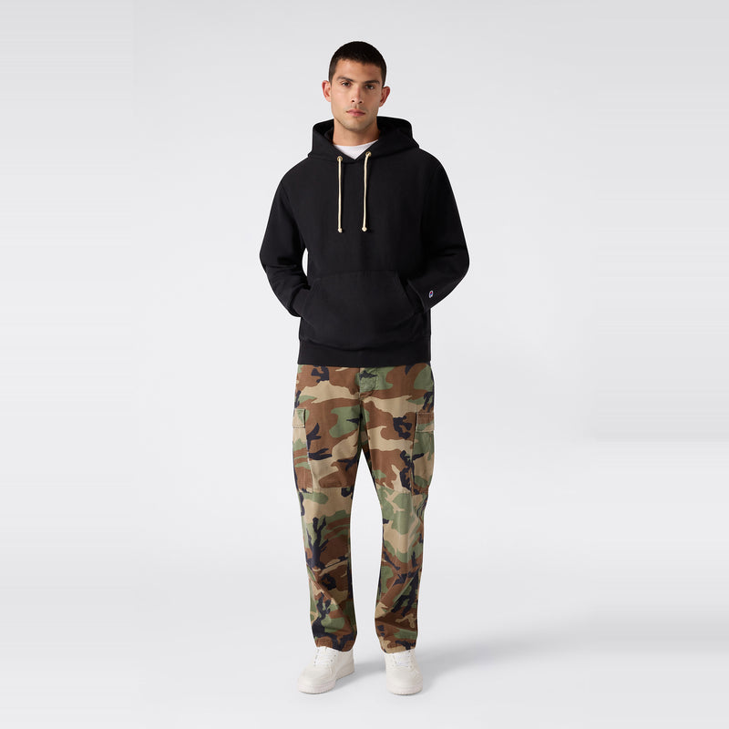 A person stands facing forward against a plain light gray background, wearing a black Champion Men’s Relaxed Fit Hooded Sweatshirt made from heavyweight fleece, paired with camouflage pants and white sneakers.