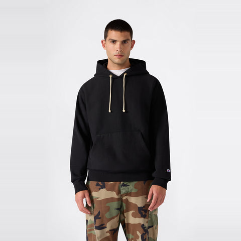 A person wearing the Champion Men’s Relaxed Fit Hooded Sweatshirt in black with beige drawstrings and camouflage pants stands against a plain white background.