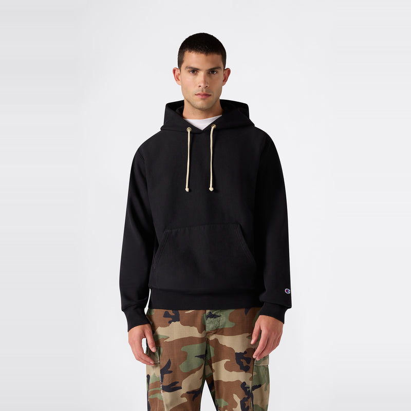 A person wearing the Champion Men’s Relaxed Fit Hooded Sweatshirt in black with beige drawstrings and camouflage pants stands against a plain white background.