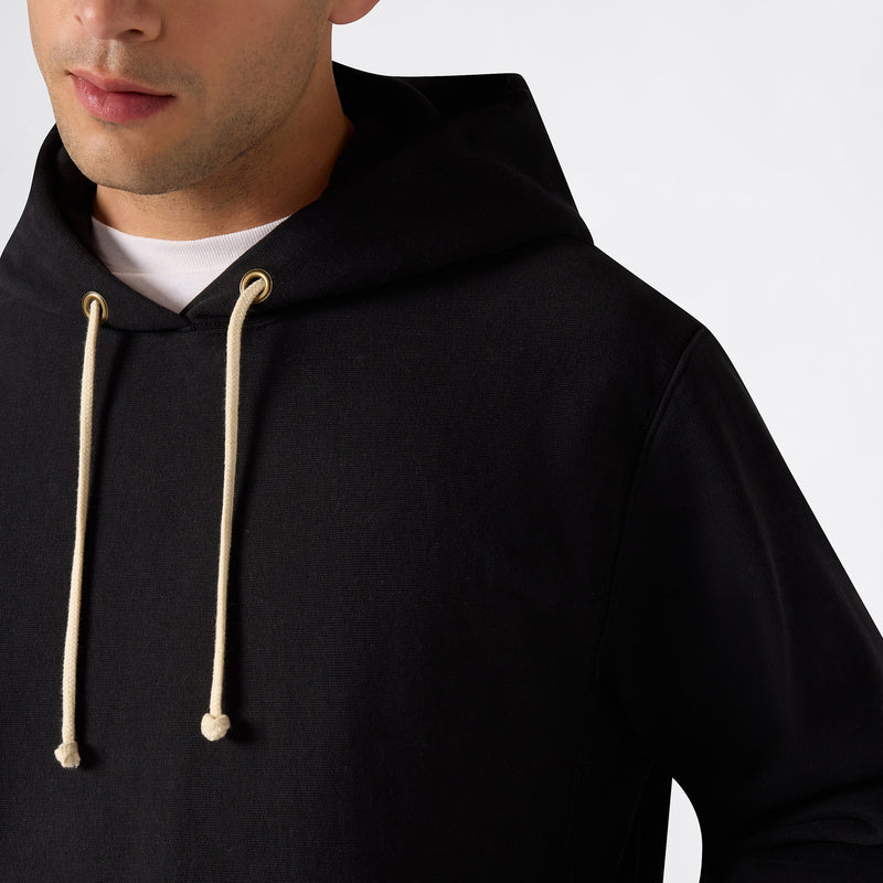 A person wearing the Champion Men’s Relaxed Fit Hooded Sweatshirt, featuring black fabric with cream-colored drawstrings and a white shirt underneath. Only the lower half of their face and upper body are visible against a plain, light background.