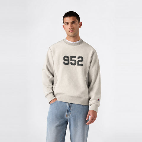 A person wearing a light gray Champion Men’s Knitted Crewneck Sweatshirt (Reversible + Vintage Wash) with the number 952 on the front, paired with light blue jeans, stands against a plain white background.