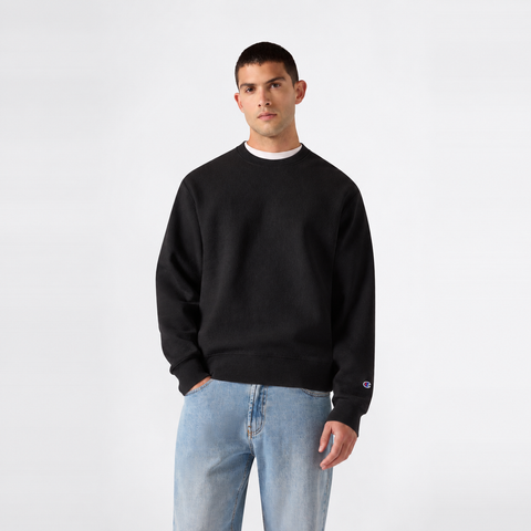 A man stands against a plain white background, wearing the Champion Men’s Knitted Crewneck Sweatshirt in black over a white shirt and light blue jeans, with one hand in his pocket. This classic sweatshirt showcases the timeless style that Champion is known for.
