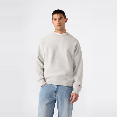 A person is wearing a Champion Men’s Relaxed Fit Crewneck Sweatshirt in plain light gray and light blue jeans, standing against a plain white background.