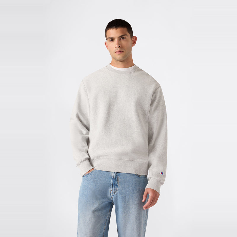 A person is wearing a Champion Men’s Relaxed Fit Crewneck Sweatshirt in plain light gray and light blue jeans, standing against a plain white background.