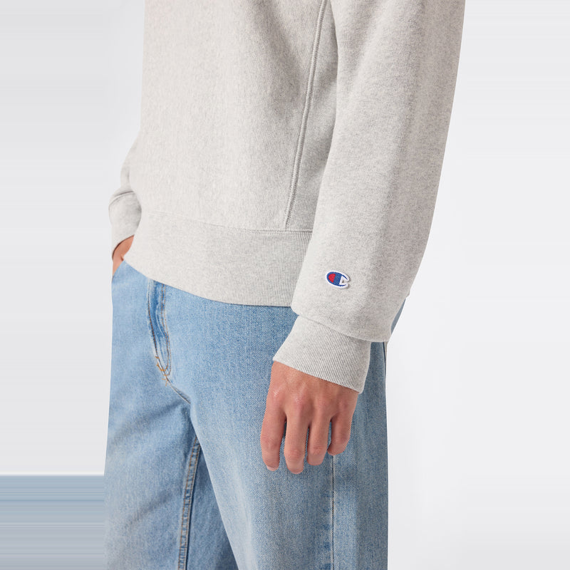 A person wearing light blue jeans and a light gray Champion Men’s Relaxed Fit Crewneck Sweatshirt, featuring the brand’s signature small logo on the sleeve, stands against a plain background. Made by Champion, this men’s sweatshirt is designed in a relaxed fit from heavyweight fleece for enduring comfort and classic style.