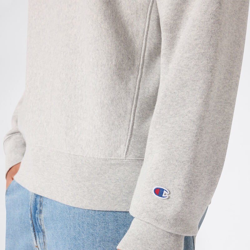 A person wearing the Champion Men’s Relaxed Fit Crewneck Sweatshirt in light gray, made from heavyweight fleece and featuring the signature small Champion logo on the sleeve, paired with light blue jeans and standing with one hand in their pocket.