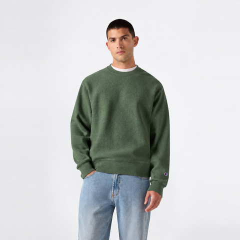 A person wearing the Champion Men’s Relaxed Fit Crewneck Sweatshirt in green and light blue jeans stands against a plain white background, looking at the camera with a neutral expression.