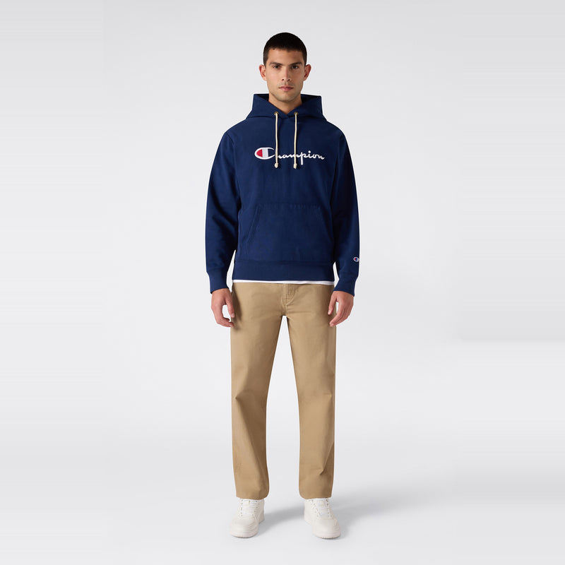 A man faces forward wearing a navy blue Champion Men’s Knitted Hooded Sweatshirt with an embroidered logo, crafted from heavyweight fleece, paired with khaki pants and white sneakers against a plain light background.