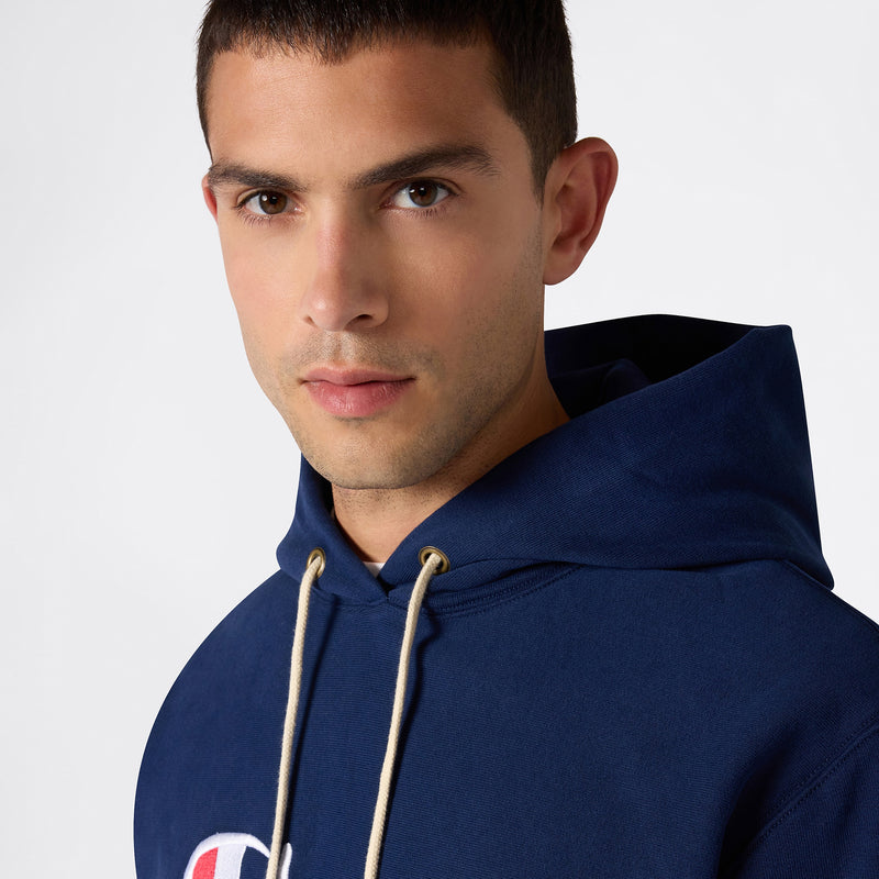 A young man with short dark hair wears a navy blue Champion Men’s Knitted Hooded Sweatshirt (Embroidered Logo) featuring cream drawstrings. Facing the camera with a neutral expression against a plain, light background, he showcases the heavyweight fleece construction of this Champion hoodie.