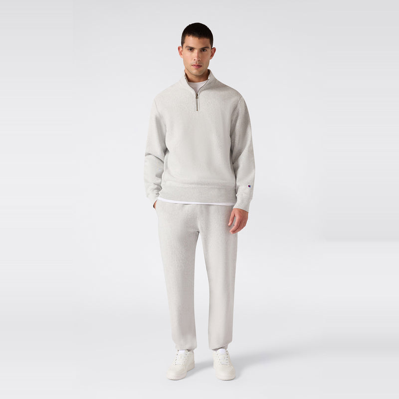 A man stands facing forward, wearing a light gray Reverse Weave quarter-zip sweatshirt and Champion Men’s Knitted Elastic Cuff Pants with white sneakers against a plain white background.