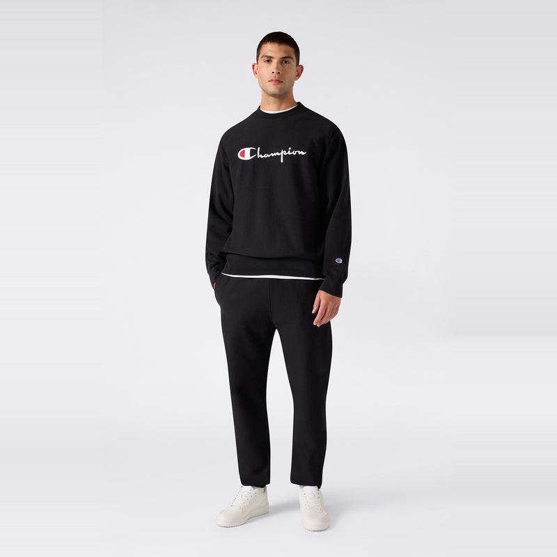A person stands against a plain background wearing a black Champion sweatshirt, Champion Men’s Knitted Elastic Cuff Pants, and white sneakers.