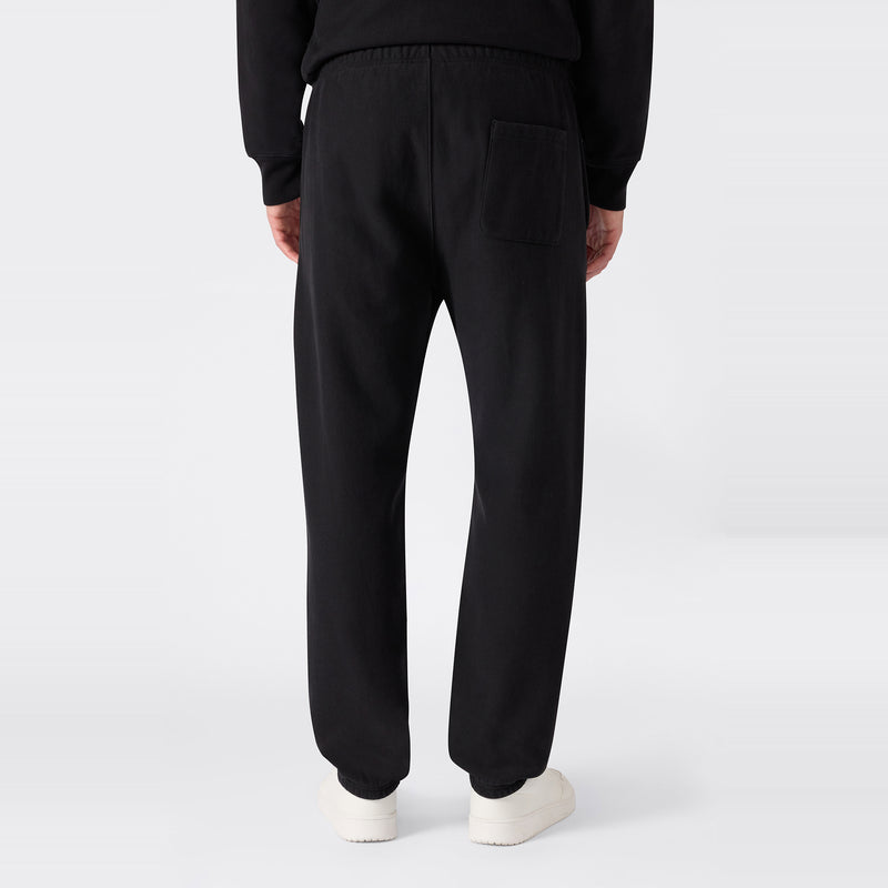A person is shown from the back against a plain light background, wearing black Champion Men’s Knitted Elastic Cuff Pants—designed in a classic sportswear fleece style with an elastic waistband and single back pocket—and white shoes.