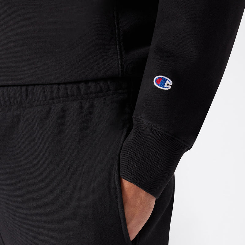 A person wearing black Champion Men’s Knitted Elastic Cuff Pants paired with a matching black sweatshirt, both featuring the iconic Champion logo embroidered on the sleeve, with one hand casually tucked into the pocket.