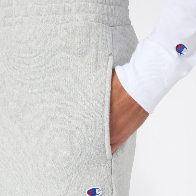 Close-up of a person wearing light gray Champion Men’s Knitted Wide Leg Pants paired with a white long-sleeve shirt, both displaying the Champion logo. Made from heavyweight fleece, the trousers combine comfort and style with their wide-leg fit, as the person’s hand rests casually in the pants pocket.