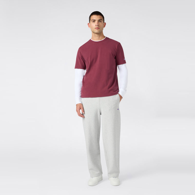 A person stands against a plain light background, wearing a maroon T-shirt layered over a white long-sleeve shirt, Champion Men’s Knitted Wide Leg Pants in gray, and white shoes.