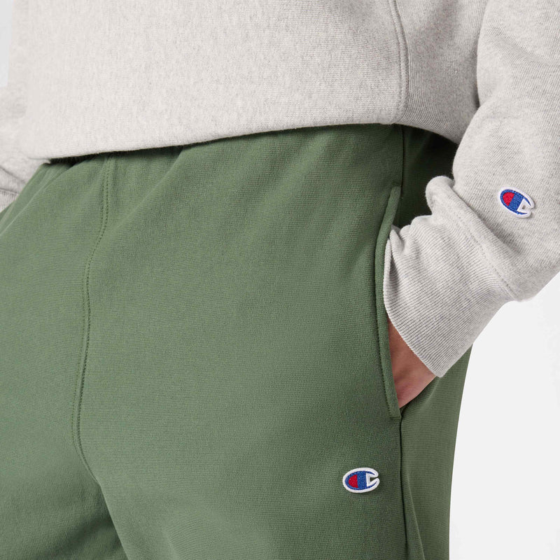 A person wearing an off-white sweatshirt and green Champion Men’s Knitted Wide Leg Pants, both featuring a small embroidered Champion logo, with their hand in the pocket.