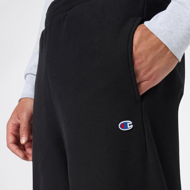 Close-up of a person wearing Champion Men’s Knitted Wide Leg Pants in black, featuring a visible logo patch and hand resting in the side pocket, paired with a light gray long-sleeved shirt against a plain white background.