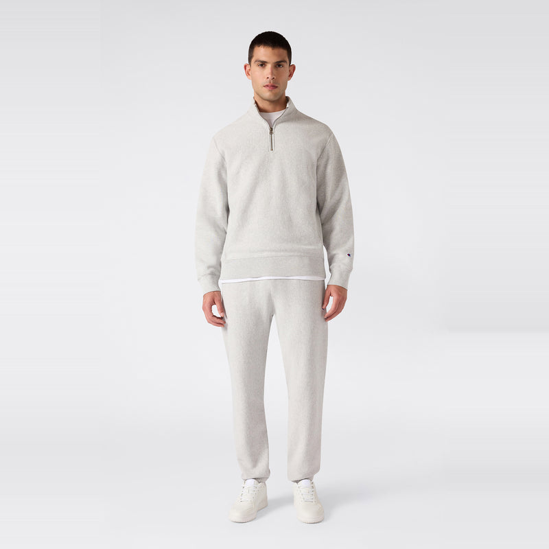 A man stands facing forward wearing a light gray Champion Men’s Knitted Half Zip Sweatshirt, matching sweatpants, a white undershirt, and white sneakers against a plain white background.
