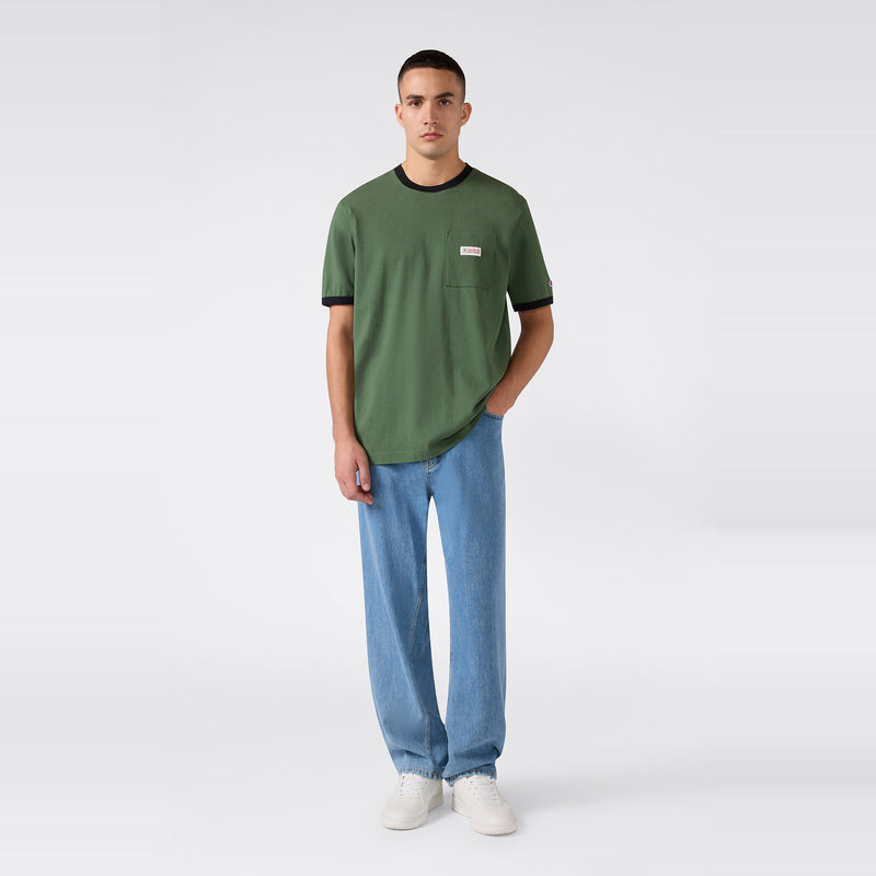 A person stands against a light, plain background wearing the Champion Men’s Knitted Ringer T-Shirt with black trim, paired with light blue jeans and white sneakers. One hand is in their pocket as they look toward the camera.