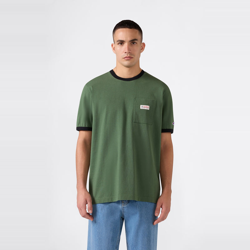 A person stands against a plain background, wearing a Champion Men’s Knitted Ringer T-Shirt in green with black trim and a chest pocket, paired with light blue jeans.