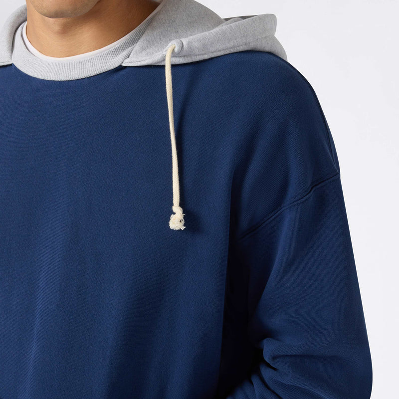 A person shown from the shoulders down against a plain background wears the Champion Men’s Knitted Hooded Sweatshirt (Vintage Wash) in blue, featuring a gray hood, white drawstring, and crafted from soft Reverse Weave fleece.