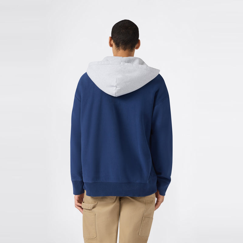 A person stands facing away, wearing a blue Men’s Knitted Hooded Sweatshirt (Vintage Wash) by Champion with a light gray hood and tan cargo pants, set against a plain white background.
