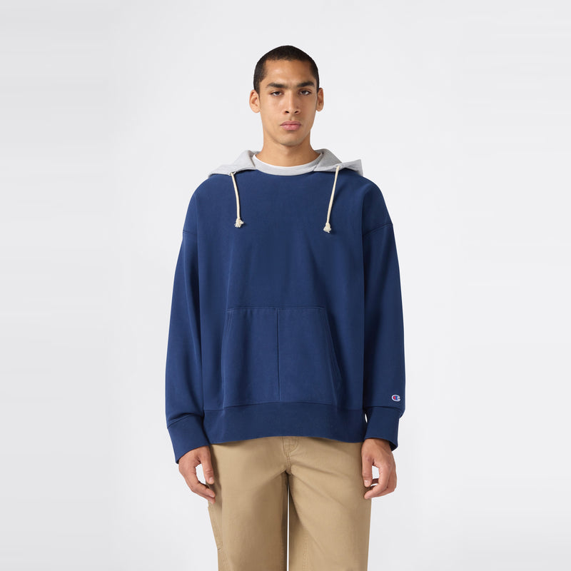 A person stands facing forward against a plain white background, wearing a blue Champion Men’s Knitted Hooded Sweatshirt (Vintage Wash) featuring light gray hood lining and drawstrings, paired with tan pants and a neutral expression.