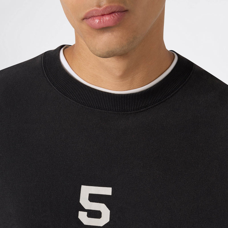 Here’s a revised version of your sentence using the provided product data: A close-up of a person wearing the Champion Men’s Knitted Crewneck Sweatshirt (Reversible + Vintage Wash) in black, featuring a white number 5 and layered over a white crew-neck shirt. Only the lower half of their face and upper torso are visible, highlighting the classic Reverse Weave fleece texture.