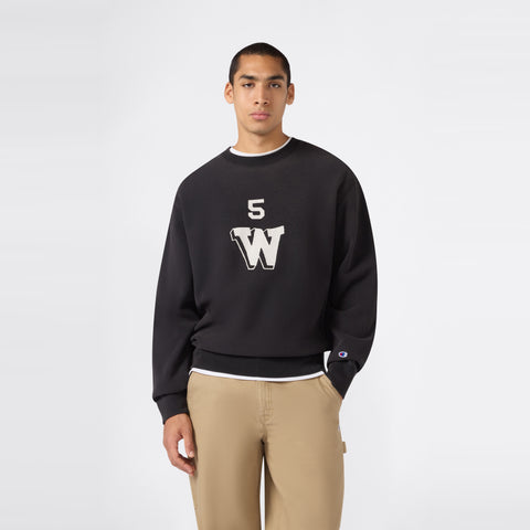A person wearing the Champion Men’s Knitted Crewneck Sweatshirt (Reversible + Vintage Wash) in black, featuring a white 5W logo, pairs it with tan pants while standing against a plain white background.
