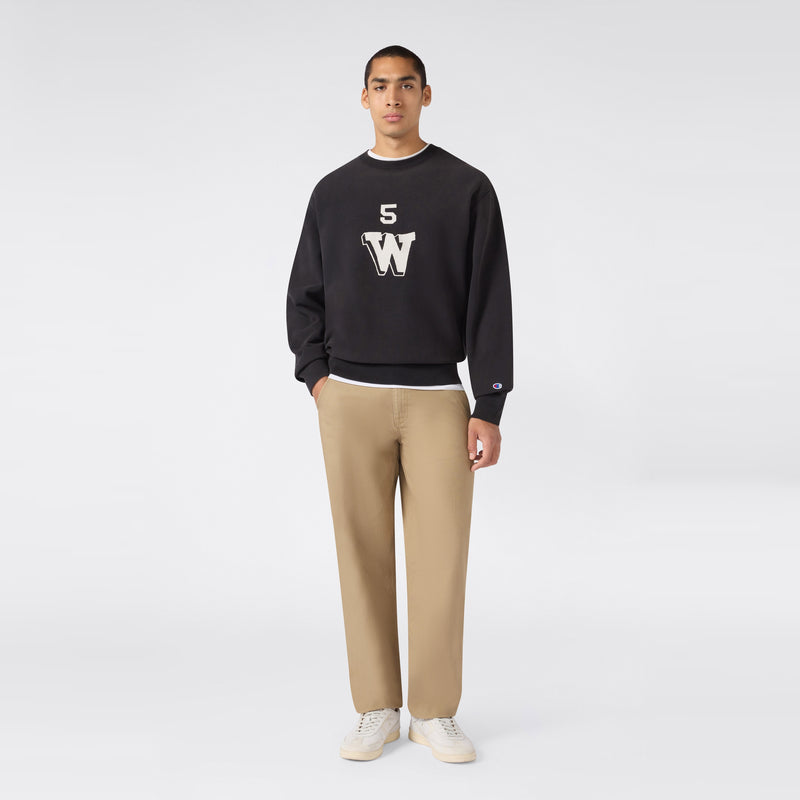 A person stands facing forward against a plain light gray background, wearing a black Champion Men’s Knitted Crewneck Sweatshirt (Reversible + Vintage Wash) featuring “5 W” lettering—crafted from signature Reverse Weave fleece for extra durability—paired with beige pants and white sneakers.