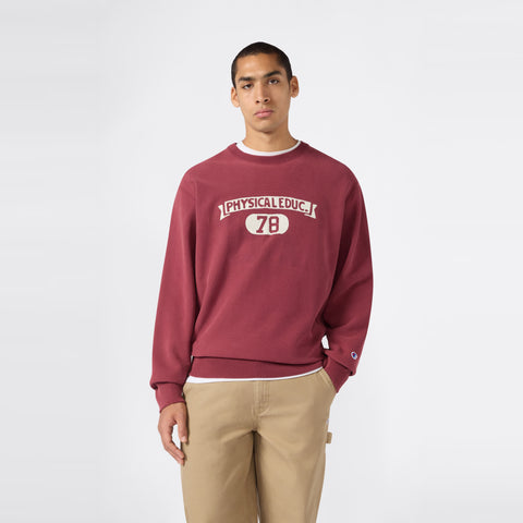 A man stands in front of a plain white background wearing the Champion Men’s Knitted Crewneck Sweatshirt (Reversible + Vintage Wash) in maroon, featuring “PHYSICAL EDUC. 78” on the front, paired with khaki pants.