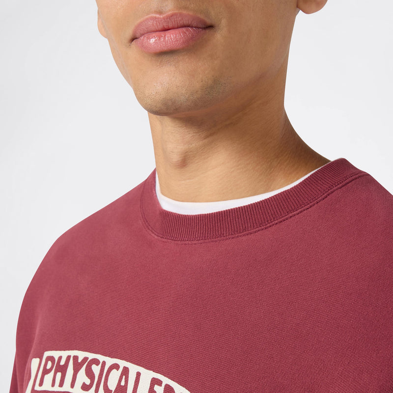 A close-up of a person wearing the Champion Men’s Knitted Crewneck Sweatshirt (Reversible + Vintage Wash) in maroon with white lettering over a white undershirt. Only the lower half of their face and upper chest are visible.