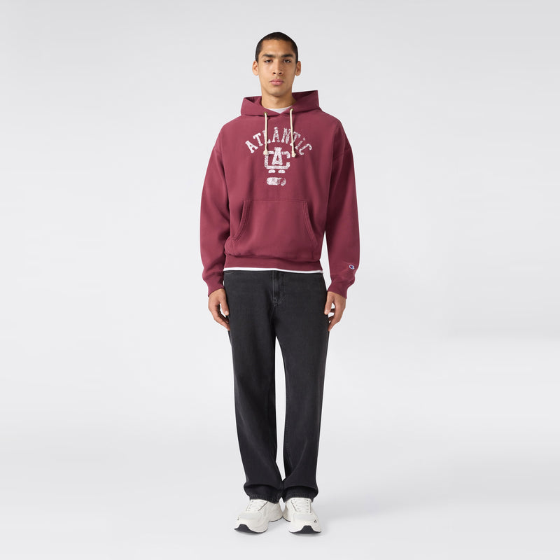 A person stands facing forward, wearing the Men’s Knitted Hooded Sweatshirt by Champion in maroon with ATLANTIC text—crafted from heavyweight fleece—and pairs it with black pants and white sneakers, set against a plain light background.