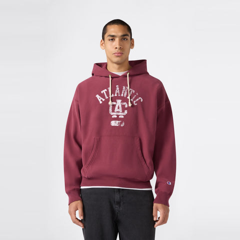 A person wearing a maroon Champion Men’s Knitted Hooded Sweatshirt featuring “ATLANTIC” and a faded graphic stands against a plain light background, looking directly at the camera with hands by their sides. This heavyweight fleece hoodie seamlessly blends comfort and timeless style for an effortless look.