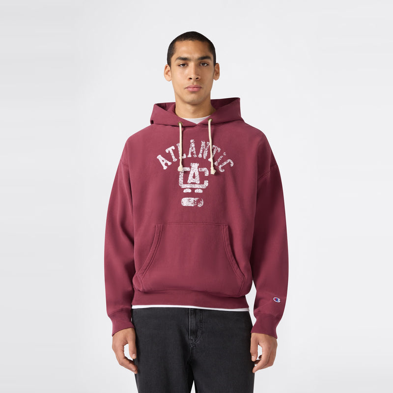 A person wearing a maroon Champion Men’s Knitted Hooded Sweatshirt featuring “ATLANTIC” and a faded graphic stands against a plain light background, looking directly at the camera with hands by their sides. This heavyweight fleece hoodie seamlessly blends comfort and timeless style for an effortless look.
